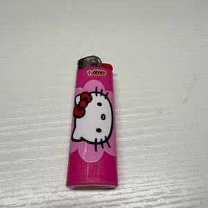 以下が含まれることがあります： ピンク色のBICライター、ハローキティのデザイン。ライターは、赤いリボンと黒いディテールが付いた白いハローキティの顔が特徴です。ライターは白い表面にあります。