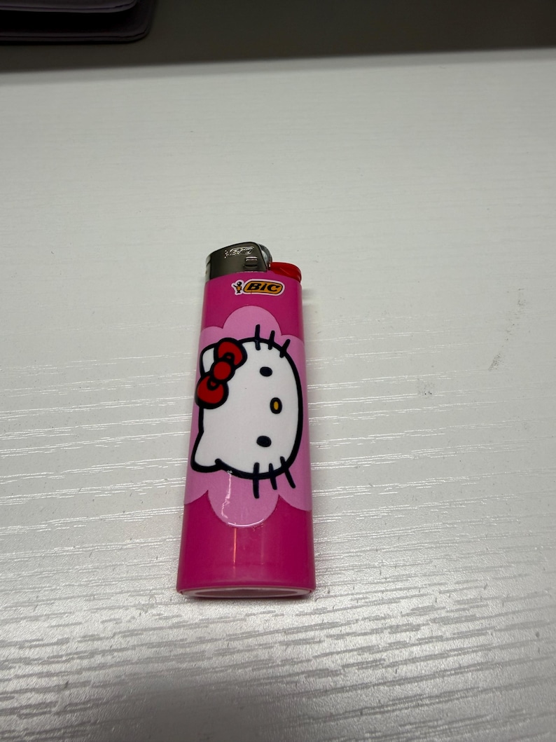 以下が含まれることがあります： ピンク色のBICライター、ハローキティのデザイン。ライターは、赤いリボンと黒いディテールが付いた白いハローキティの顔が特徴です。ライターは白い表面にあります。