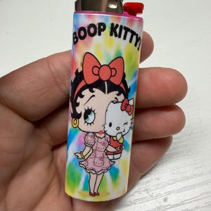 May include: A Bic lighter featuring a tie-dye background with a colorful graphic of Betty Boop holding a Hello Kitty doll. The text BOOP KITTY! is printed in black at the top. The lighter has a metallic silver top and a red ignition button.