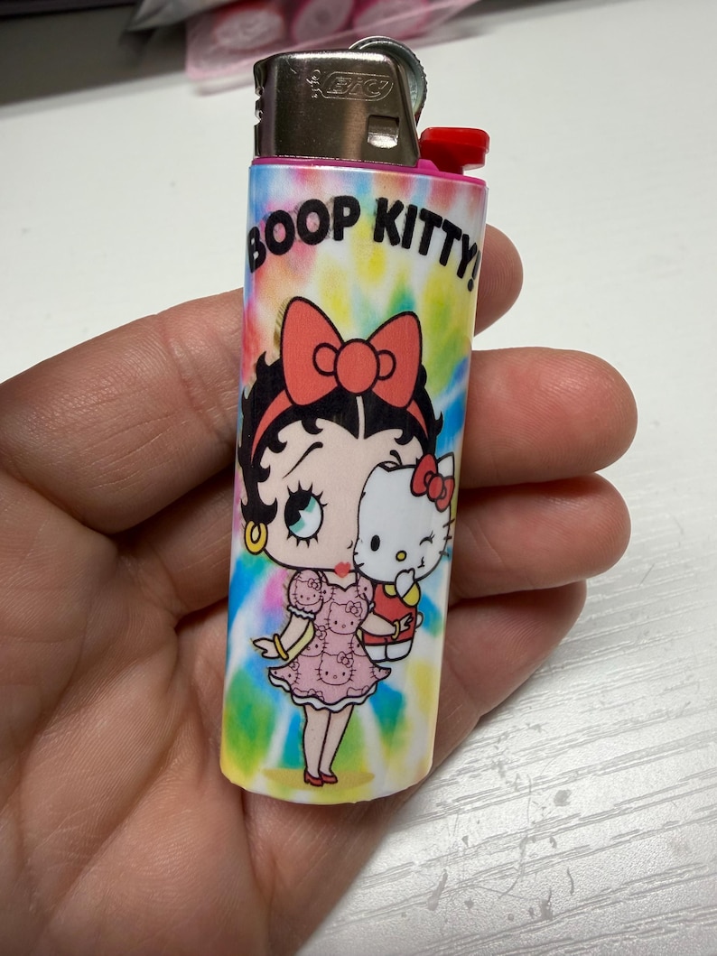 May include: A Bic lighter featuring a tie-dye background with a colorful graphic of Betty Boop holding a Hello Kitty doll. The text BOOP KITTY! is printed in black at the top. The lighter has a metallic silver top and a red ignition button.