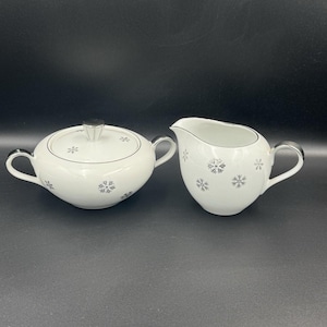 May include: A white porcelain sugar bowl with a lid and a creamer. Both pieces have silver rims and are decorated with grey snowflake designs. The sugar bowl has two handles, and the creamer has one handle and a spout.