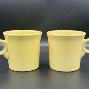 Könnte beinhalten: Zwei hellgelbe Keramikbecher mit abgerundeten Griffen. Die Becher haben ein klassisches Design mit leicht ausgestelltem Rand und glatter, glänzender Oberfläche. Die Becher stehen nebeneinander vor einem dunklen Hintergrund.