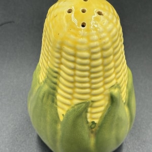 May include: A ceramic salt or pepper shaker shaped like an ear of corn. The top portion features yellow kernels with five small holes for dispensing, while the base is surrounded by textured green husks. The surface has a glossy, smooth finish.