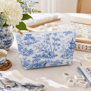 May include: A zipped pouch featuring a blue and white toile-style pattern depicting a landscape with bamboo, a bridge, and a pagoda. The design includes small mahjong tile illustrations. The pouch has a gold-toned metal zip pull.