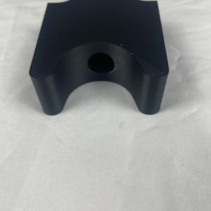 Puede incluir: Un objeto negro y rectangular con una parte superior curvada y un agujero en el centro. El objeto tiene dos patas curvas en la parte inferior. El artículo está sobre una superficie blanca.