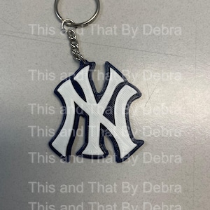 Puede incluir: Un llavero con el logotipo de los Yankees de Nueva York. El logotipo es un diseño "NY" blanco con un contorno azul marino. El llavero incluye una cadena y un llavero plateados. El texto "This and That By Debra" está en la marca de agua en el fondo.