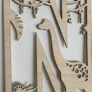 May include: Wooden letter "N" with a dinosaur theme. The letter is cut out of light-colored wood and features a long-necked dinosaur, a triceratops, and jungle foliage. The design is suitable for nursery decor or children's room.