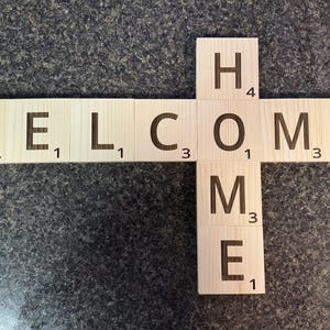 May include: Wooden tile sign spelling "WELCOME HOME" in dark brown letters. The letters are arranged in a cross shape, with the word "WELCOME" horizontally and "HOME" vertically. Each tile has a point value.