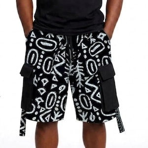 Puede incluir: Pantalones cortos tipo cargo de color negro con un estampado gráfico abstracto en blanco de formas geométricas y líneas. Los pantalones incluyen dos grandes bolsillos laterales con solapa y un cordón negro en la cintura, junto a una camiseta negra lisa.