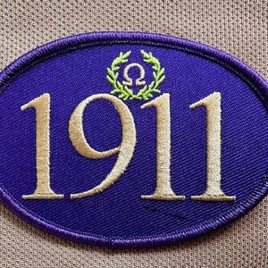 Peut inclure: Écusson brodé ovale violet avec une bordure violette. Il affiche l'année "1911" en crème, avec un petit emblème au-dessus du "9". Le fond est un tissu texturé de couleur beige.