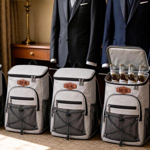 May include: Four light gray insulated cooler backpacks with black zippers and mesh front pockets. Each bag features a brown leather-like patch with custom initials. One backpack is open, revealing four glass beer bottles inside the main compartment.