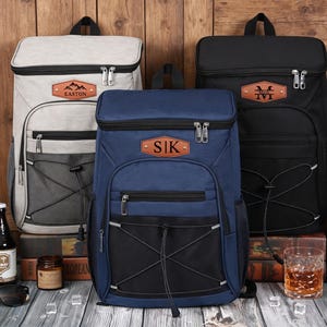 May include: Three rectangular backpacks in light gray, navy blue, and black, each featuring a top handle, a front zippered pocket, a black mesh bungee cord system, and a brown leather-like patch with personalized text including Easton, S|K, and Mark.