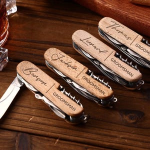 Puede incluir: Cuatro multiherramientas de madera con nombres personalizados y la palabra "Groomsman" grabada. Cada herramienta tiene una hoja plateada y varios implementos. Las herramientas están dispuestas sobre una superficie de madera oscura.