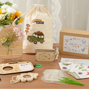 Puede incluir: Una colección de artículos con temática de la naturaleza, que incluye una pantalla de madera con flores, una bolsa con cordón con un gráfico de jeep, una caja etiquetada como "Plant Explorer Kit" y un artículo de madera con forma de cámara. Otros artículos incluyen una lupa y pinzas.