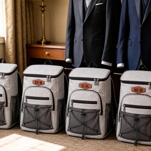 May include: Four light gray backpacks with black trim and mesh side pockets. Each bag features a brown leather-like patch on the front with unique initials: N|S, D|R, S|K, and M|B. The backpacks have a top handle, a front zippered pocket, and a black bungee cord system.
