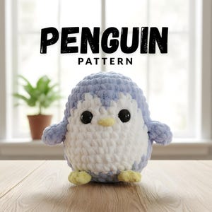 Op de afbeelding: Een gehaakt pinguïnspeelgoed in blauw en wit, met zwarte ogen en een gele snavel en voeten. Het woord "PENGUIN" staat in grote, vette, zwarte letters boven het woord "PATTERN". De pinguïn staat op een houten oppervlak.
