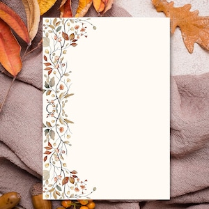 May include: A cream-colored stationery card with a floral border of autumn leaves and berries. The card is surrounded by fall foliage, including orange and yellow leaves, acorns, and a brown fabric.