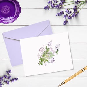 May include: A white card with a watercolor illustration of a lilac flower bouquet. The card is on a white wooden surface with a lavender sprig and a purple saucer.
