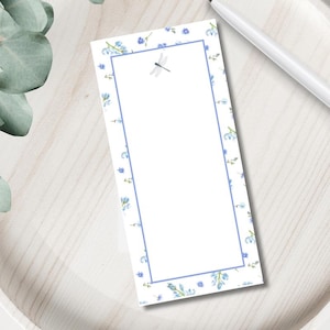 May include: A rectangular notepad with a white background, adorned with a delicate floral pattern in shades of blue. A light blue border frames the blank space, with a dragonfly illustration at the top. A white pen rests nearby.