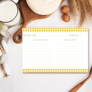 May include: A white recipe card with a yellow gingham border. The card has spaces for recipe name, ingredients, servings, and directions. The card is on a white surface with a wooden cutting board, milk, and walnuts.