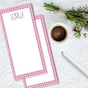 May include: Two white notepads with a pink gingham border and the initials "JMJ" in pink script. A cup of coffee, a pen, and a small bouquet of flowers and greenery are also on the white, textured surface.