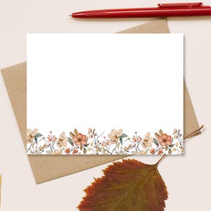 May include: A white stationery card with a floral border featuring peach and cream-colored flowers and green leaves. A red pen rests at the top, and a brown envelope is beneath the card. A brown leaf is in the foreground.
