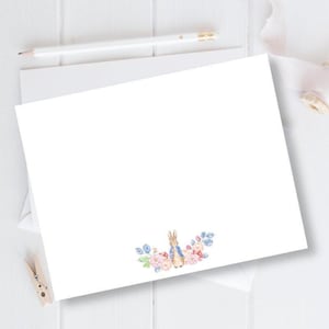 May include: A blank white card with a floral border featuring a watercolor illustration of Peter Rabbit in a blue jacket. The flowers are pink, blue, and white.