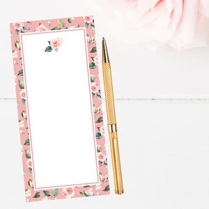 May include: A gold pen rests beside a pink floral notepad. The notepad features a white interior with a pink border and is decorated with roses and green leaves. The pen has a textured design and a gold finish, complementing the notepad's floral theme.