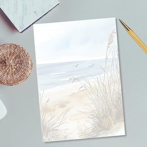 May include: A watercolor print of a beach scene with sand, sea oats, and the ocean. The artwork features a light blue sky and two birds in flight. The print is on a white paper, with a gold pen and a woven basket in the background.