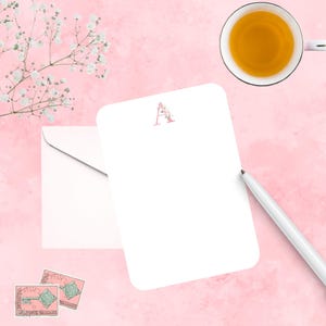 May include: A white stationery set with a floral "A" initial, a matching envelope, and a pen on a pink background. A cup of tea and two vintage postage stamps are also present. The stationery is perfect for personalized notes.