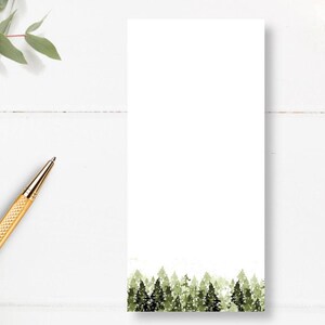 May include: A blank white notepad with a green watercolor forest design at the bottom. The notepad is on a white wooden surface with a gold pen.
