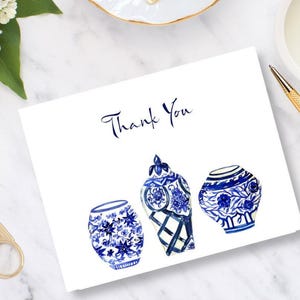 May include: A white card with the words "Thank You" in navy blue script. The card features watercolor illustrations of three blue and white decorative vases. Gold scissors, a pen, and a candle are in the background.