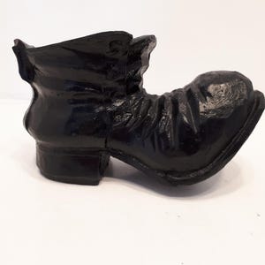 May include: A black decorative boot, possibly a planter or ornament. The boot features a rounded toe, a small heel, and a ruffled upper cuff. The surface appears glossy, indicating a lacquered or painted finish. It is positioned against a plain white backdrop.