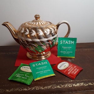 Peut inclure: Une théière dorée ornée de motifs de feuilles blanches et d'un motif floral rouge et vert. Des sachets de thé Stash et Twinings, dont de la menthe poivrée et du English Breakfast, sont disposés autour.