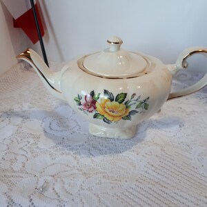 May include: Cream ceramic teapot with a floral design of yellow and pink roses. The teapot has gold trim on the spout, handle, and lid. The teapot is on a white lace tablecloth. A charming tea accessory.