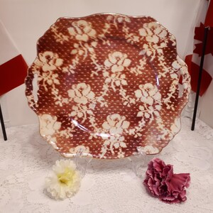 May include: A decorative plate with a scalloped edge, featuring a brown background with a white floral pattern. The plate is displayed on a clear stand, with two Canadian flags in the background. Two flowers are placed near the plate.