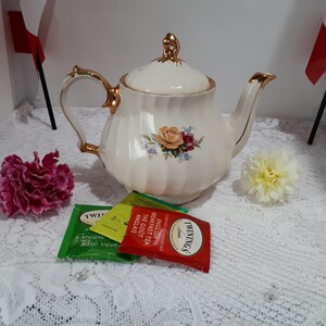 May include: A cream-coloured porcelain teapot with gold trim, a floral design, and a matching lid. Tea bags and flowers are arranged around the teapot. The teapot is on a white lace tablecloth.