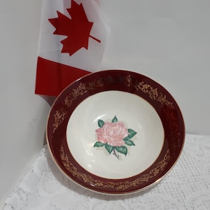 May include: A decorative bowl with a claret rim and a cream-coloured centre. The centre features a pink rose with green leaves. The rim is adorned with gold filigree. A Canadian flag is in the background.