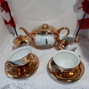 May include: A gold-plated tea set with white and blue detailing, comprising a teapot, sugar bowl, creamer jug, and two teacups with saucers. The set is displayed on a white lace tablecloth, with a decorative design.