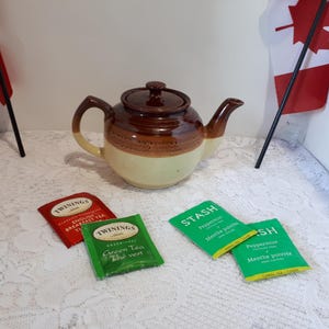 May include: A brown and cream ceramic teapot with a lid, handle, and spout. Twinings and Stash tea bags are in front of the teapot. Two Canadian flags are in the background.