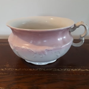 May include: A vintage ceramic chamber pot in a pale pink and white colour scheme. The pot has a rounded body, a decorative handle, and embossed floral detailing. The rim and base are white, while the body transitions from pink to white.