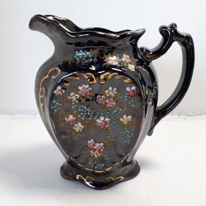 May include: Black ceramic jug with a decorative floral design. The jug features a curved handle and a spout. The floral design includes pink, white, and blue flowers with gold accents. The jug has a glossy finish.