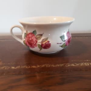 May include: A white ceramic cup with a handle, decorated with red and pink rose floral designs and green leaves. The cup is on a dark brown wooden surface.