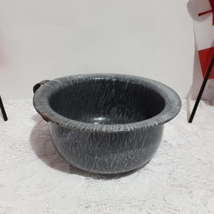 May include: A grey and white speckled enamelware bowl with a handle. The bowl has a wide rim and a rounded shape. The bowl is on a white lace tablecloth. Two Canadian flags are in the background.
