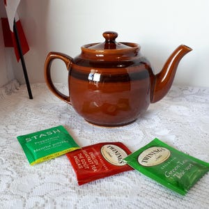 May include: A brown ceramic teapot with a lid, handle, and spout, sits on a white lace tablecloth. Three tea packets are in front of the teapot. The tea packets are green, red, and green, with text on them.