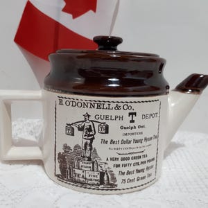 May include: A vintage ceramic teapot with a brown lid and spout, and a white body. The teapot has a handle and a spout. The body of the teapot has a label with text and an illustration of a person carrying boxes. A Canadian flag is in the background.