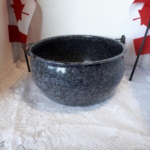 May include: A speckled grey granite cooking pot with two metal handles. The pot is round and sits on a white lace tablecloth. Two Canadian flags are in the background.