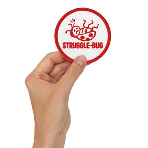 Pode incluir: Um patch redondo branco com uma borda vermelha e as palavras "STRUGGLE-BUG" em vermelho. O patch apresenta um desenho de joaninha vermelha de desenho animado. O patch é segurado por uma mão em um fundo branco.