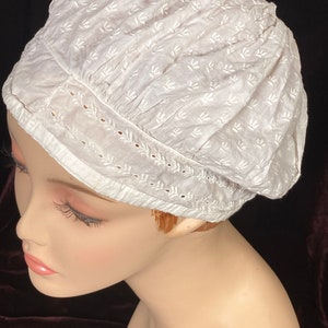 May include: White cotton bonnet with a delicate floral pattern and lace trim. The bonnet is gathered at the top and has a rounded shape.
