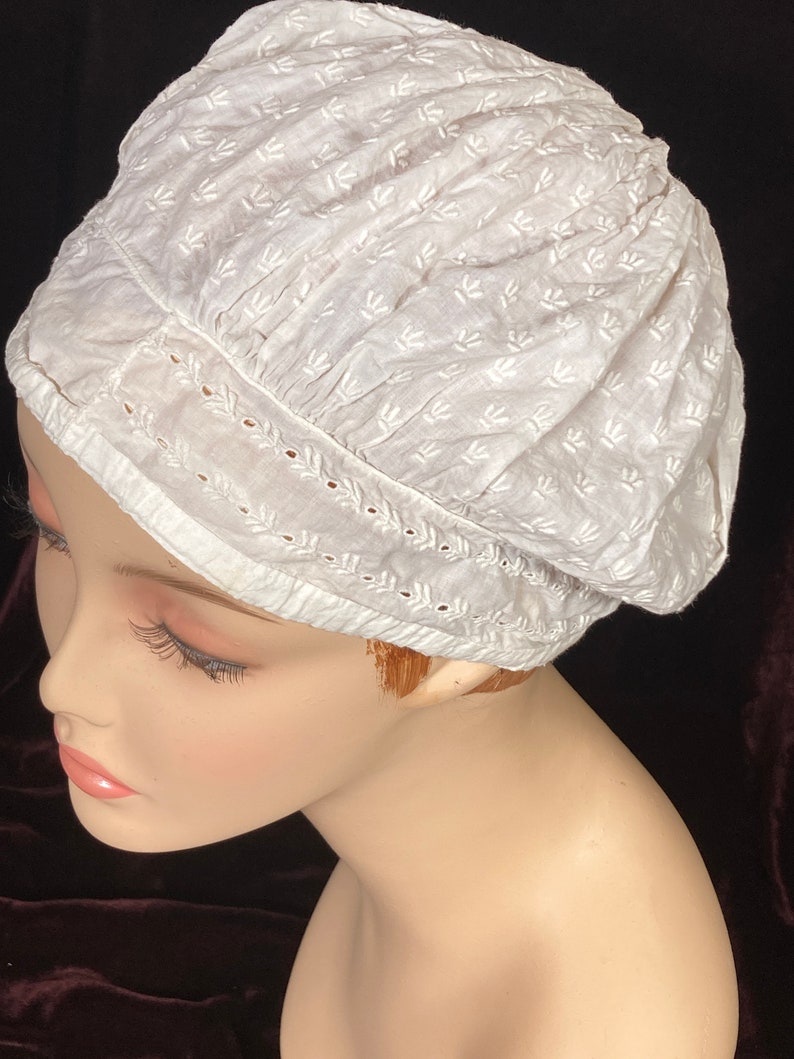 May include: White cotton bonnet with a delicate floral pattern and lace trim. The bonnet is gathered at the top and has a rounded shape.
